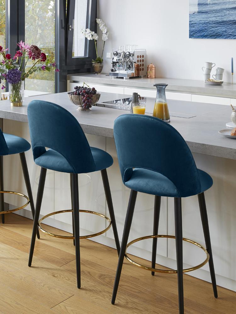 Aksominė mėlyna baro kėdė Rachel su juodomis matinėmis kojomis ir auksine atrama, pastatyta modernioje virtuvėje prie baro stalo.