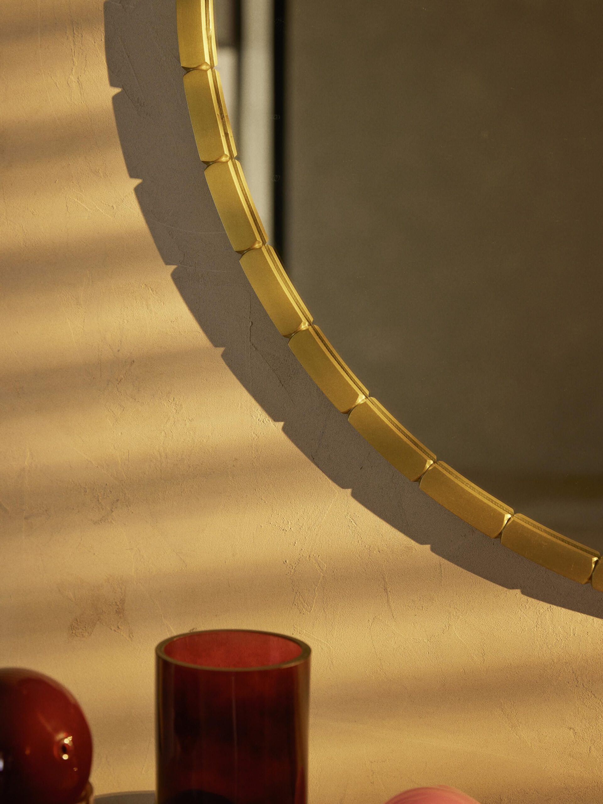 Auksinio rėmo apvalus veidrodis Alaia (Ø 82 cm) šiltame apšvietime, su subtiliomis tekstūromis ir dekoratyviniais elementais fone.