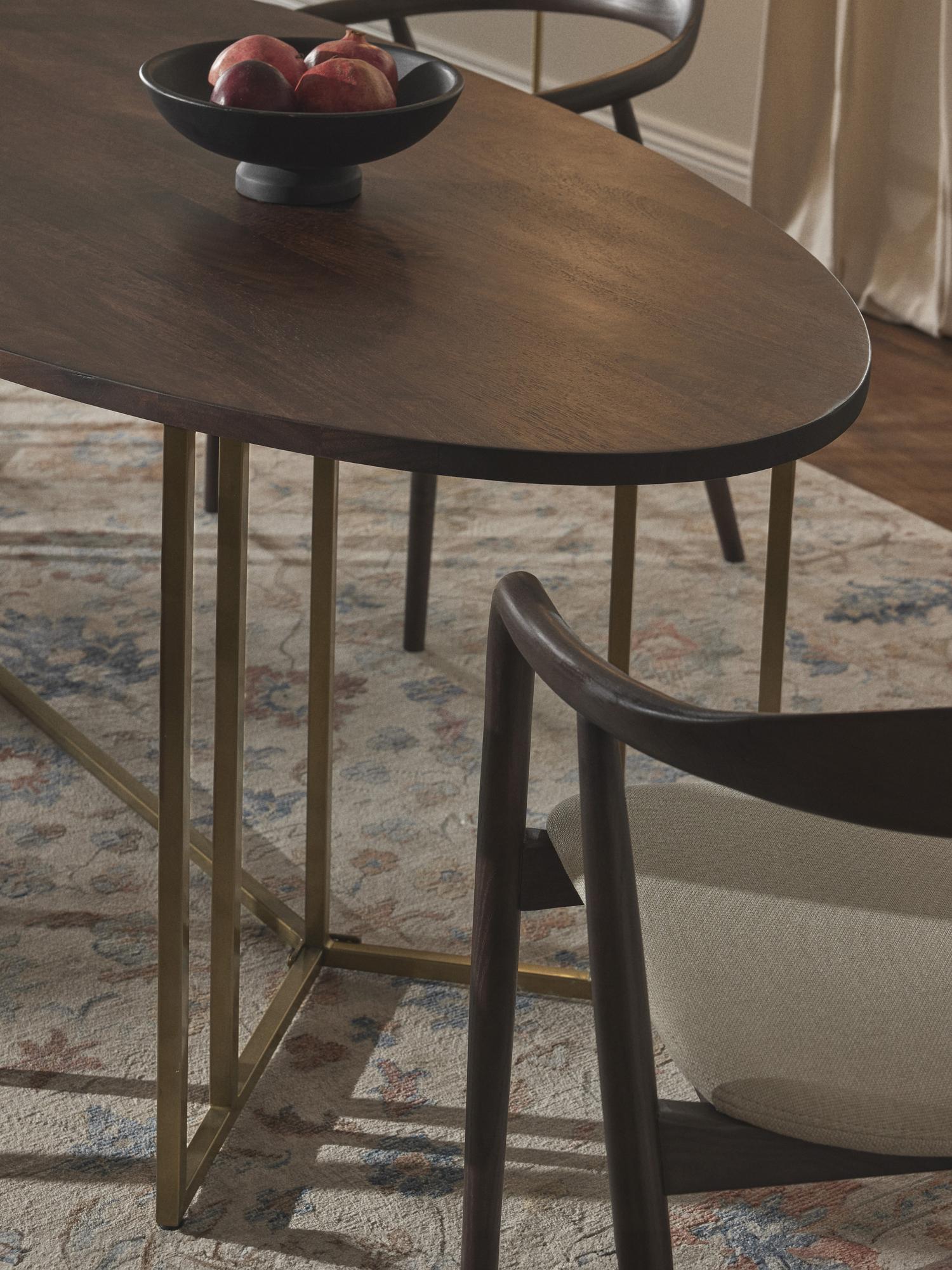 Ovalus pietų stalas Luca iš mango medienos su tamsaus medžio stalviršiu ir auksiniu metaliniu rėmu, pastatytas ant raštuoto kilimo.