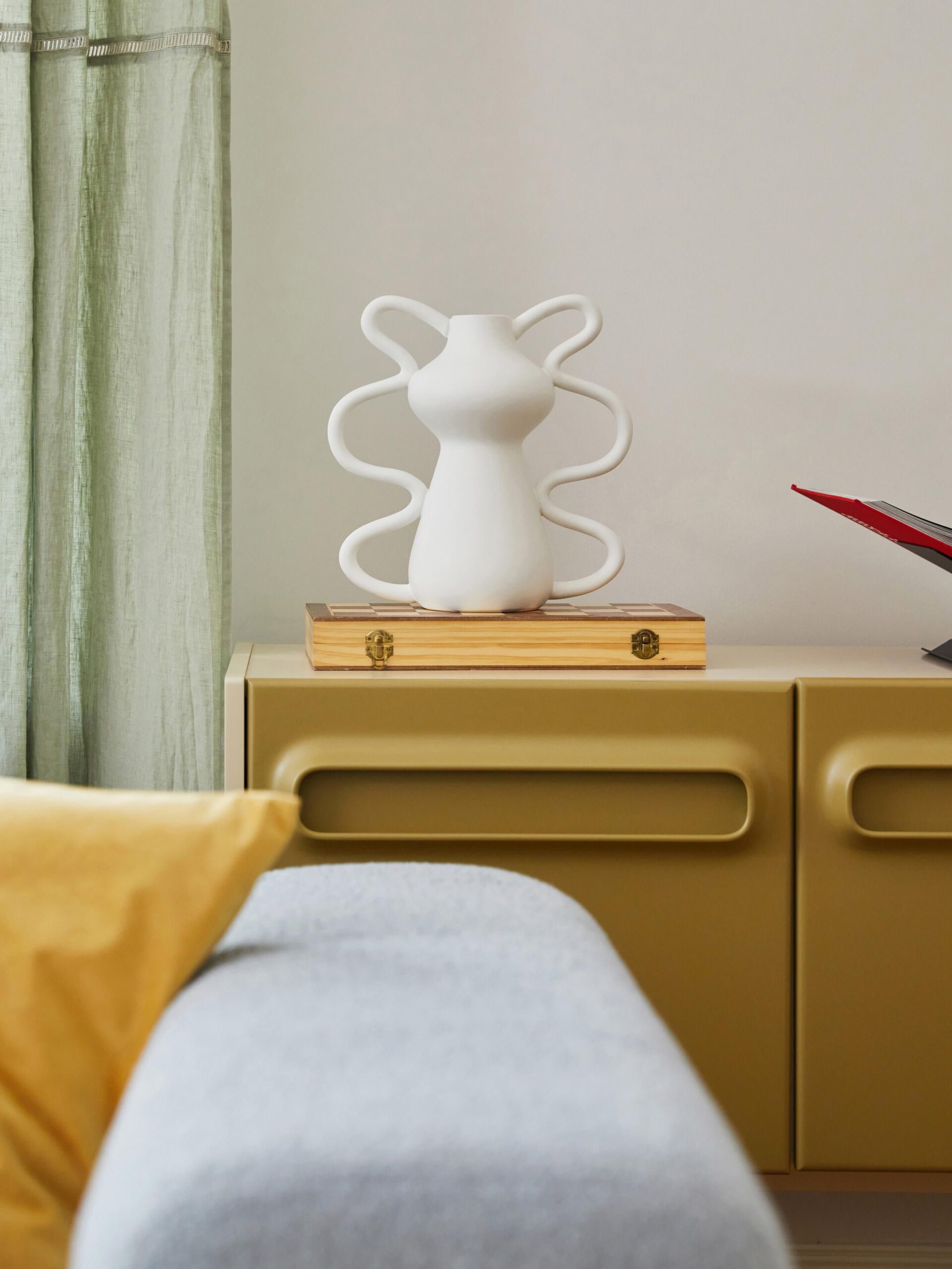 Organinės formos balta matinė vaza Luvi, 32 cm, pastatyta ant medinės lentelės, moderniame interjere su šviesiai žaliomis užuolaidomis ir minimalistiniais baldais.